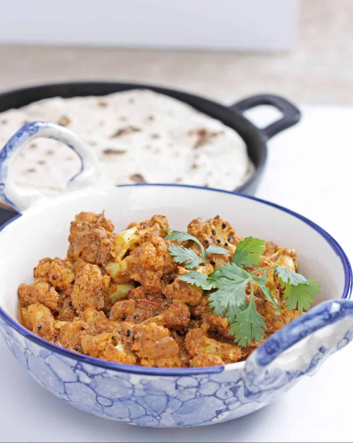 Cauliflower curry in a blue bowl garnished with cilantro and roti in the background
